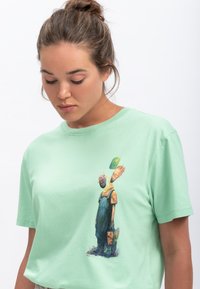T-shirt en coton vert menthe avec un imprimé graphique de deux personnages stylisés portant des masques, présenté dans un design décontracté.