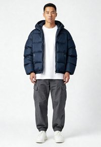 Giacca a vento imbottita blu navy con cappuccio, caratterizzata da un design trapuntato e polsini elasticizzati, abbinata a pantaloni cargo grigi e sneakers bianche.