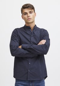 Camisa de botones azul marino con cuello mao, mangas largas y placket frontal. La tela tiene una textura suave y una forma ajustada.