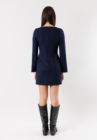 Abito blu navy con maniche lunghe, design aderente e chiusura con zip sul retro. Abbinato a stivali neri in pelle al ginocchio. Texture liscia.