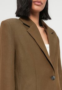 Γυναίκα που φοράει καφέ σακάκι ραμμένο στα μέτρα πάνω από λευκή μπλούζα, με σκούρα μαλλιά μήκους μέχρι τους ώμους, απεικονιζόμενη από τον λαιμό ως το μέσο του κορμού, πάνω σε απλό φόντο.