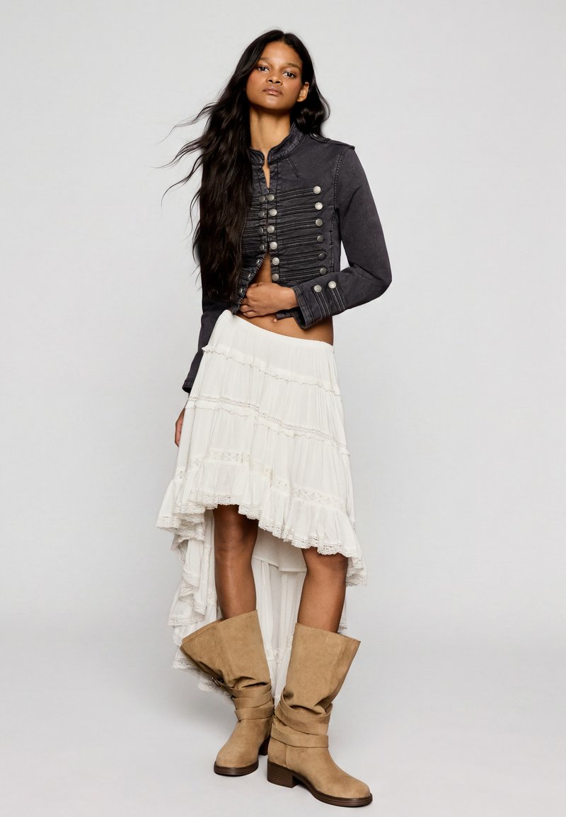 Femme aux longs cheveux portant une veste noire style militaire, une jupe asymétrique blanche et des bottes souples couleur beige, debout devant un fond uni.