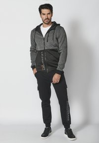 Sudadera con cremallera en tonos gris y negro, con una parte superior texturizada, una parte inferior negra sólida y capucha con cordones. Combinada con pantalones cargo negros y zapatillas deportivas.