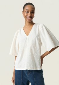 White short-sleeve top with a V-neck, textured fabric, and small embroidered dots. Loose fit, slightly cropped at the waist. Paired with dark denim.
