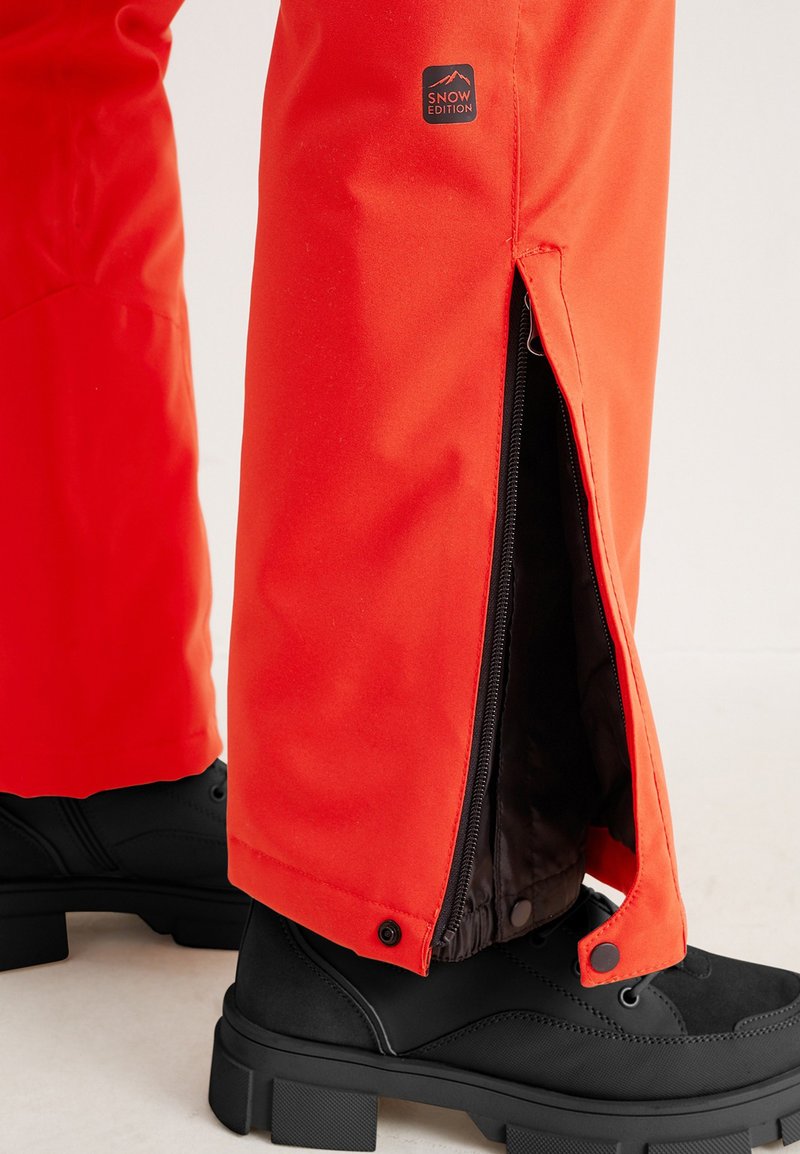 Rote wasserdichte Schneehosen mit seitlichen Reißverschlüssen, kombiniert mit schwarzen robusten Stiefeln mit texturierten Sohlen und glatten Oberflächen.