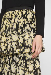 Acercamiento de la mano de una mujer sosteniendo una falda plisada con un fondo negro y patrones de margaritas amarillas y blancas, combinada con una blusa negra.