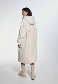 Kurtka z kapturem w miękkim, kremowym polarku o luźnym kroju, z tylną kieszenią i ściąganym dołem. W komplecie z wysokimi beżowymi botkami.