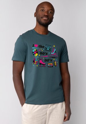 Mann trägt türkisfarbenes Shirt mit bunten Ikonen der 80er und 90er Jahre, darunter Schallplatte, Ghettoblaster, Rollschuhe, Rubik's Cube, Kassette und Mikrofon.