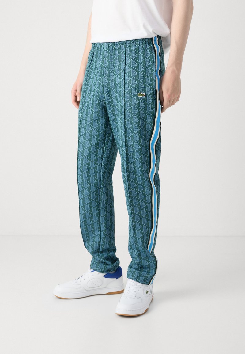 Pantalons de sport verts à motifs avec une taille élastique, des rayures latérales bleues et un logo Lacoste. Portés avec des baskets blanches.