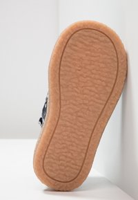 Semelle d'une chaussure bleue et blanche avec une semelle extérieure en caoutchouc texturé marron clair, reposantverticalement contre un mur et un sol blancs.