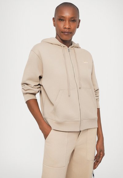 Bēšas krāsas zip-up hoodie ar lielu kapuci, priekšējām kabatām un ribotām piedurknēm. Uzkrītošs, maigs logotips uz krūšu daļas. Gluda, mīksta tekstūra.