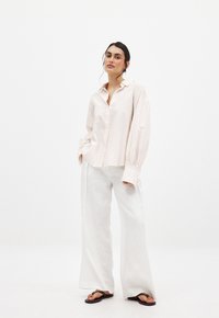 Lichtroze gestreepte overhemd met oversized mouwen, gecombineerd met losse witte linnen broeken en zwarte slippers, tegen een effen achtergrond.