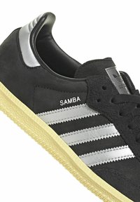 Černé semišové tenisky se stříbrnými třemi pruhy a logem. Gumová podrážka poskytuje texturovaný povrch. Elegantní design s koženým detailem na patě.