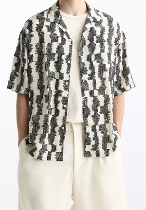Homme portant une chemise à manches courtes boutonnée avec un motif abstrait noir et blanc sur un t-shirt écru uni et un pantalon écru assorti.