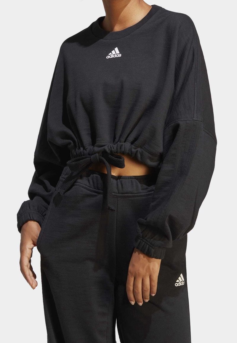 Svart kort sweatshirt med dragsko i midjan, långa ärmar och vitt Adidas-logga på bröstet. Mjuk material, avslappnad passform.