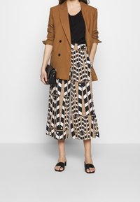 Brown double-breasted blazer over a black top, paired with a patterned midi skirt in beige, white, and black. Black slide sandals and clutch.