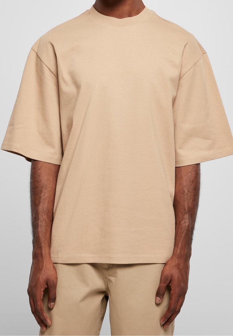 Personne portant un t-shirt beige uni oversize à manches courtes et un pantalon beige assorti sur un fond gris neutre.
