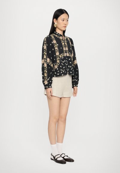 Blusa nera con stampa floreale, maniche lunghe, colletto alto e taglio cropped, abbinata a shorts beige chiari e scarpe con cinturino nere.