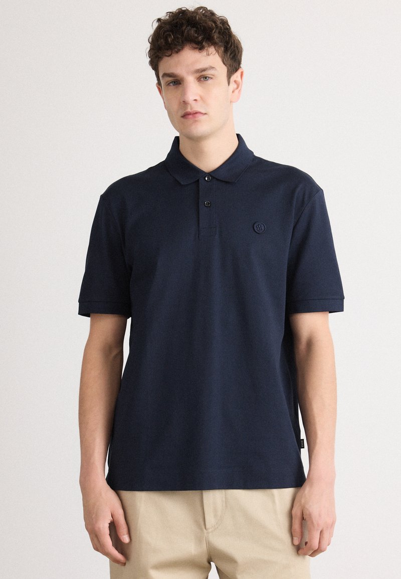 Pólo azul-escuro feito de algodão, com colarinho, pala de dois botões e um logótipo bordado no lado esquerdo do peito. Mangas curtas.