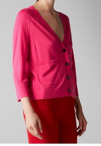 Fuchsia gebreide cardigan met een diepe V-hals, driekwart mouwen en twee voorzakken. Zwarte knopen zorgen voor contrast.