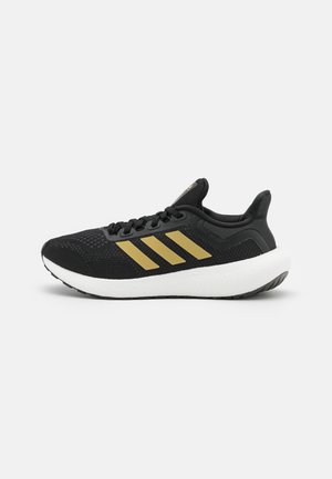 Zapatilla de running negra con rayas doradas, parte superior de malla y suela acolchada blanca y gruesa sobre un fondo blanco.