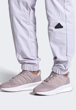 Osoba nosząca jasnofioletowe buty sportowe Adidas oraz białe spodnie Adidas z elastycznymi mankietami i małym czarnym logo Adidas na nodze.