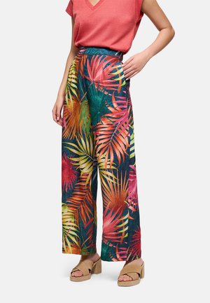 Mujer con pantalones acampanados de estampado tropical multicolor, camiseta coral de manga corta y sandalias de tacón beige, de pie con una mano en la cadera.