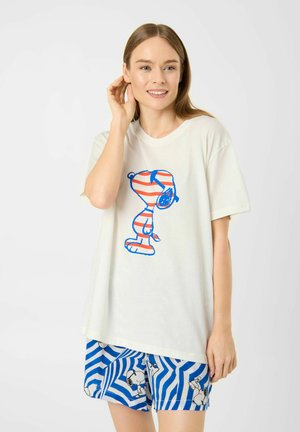 Femme portant un t-shirt blanc avec un motif Snoopy à rayures rouges et bleues et un short à rayures bleues et blanches avec motif Snoopy, souriante et se touchant l'oreille.