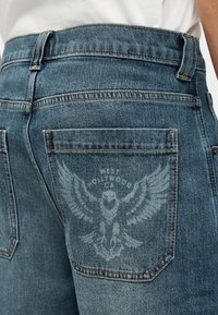 Τζιν παντελόνι με ξεβαμμένο γραφικό αετού και κείμενο στην πίσω τσέπη, φορεμένο με λευκό πουκάμισο που έχει μπει μέσα.