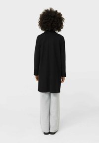 Manteau en laine noire à coupe droite, manches longues et col, associé à un pantalon de survêtement gris clair à jambes larges. Design simple et texture douce.