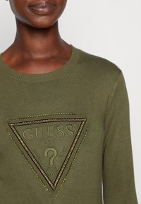 Maglione verde oliva con scollatura rotonda, caratterizzato da un logo triangolare testurizzato in beige e borchie dorate, centrato sul petto.