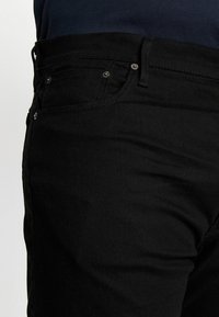 Černé džíny z denimu, které mají úzký střih, pět kapes a kontrastní kovové detaily. Tkanina má hladký, mírně strukturovaný povrch.