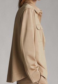 Chemise beige à manches longues en tissu lisse. Dotée d'une fermeture à boutons sur le devant, d'une poche poitrine et d'une coupe décontractée avec un détail d'épaule.