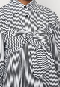 Chemise à rayures verticales noires et blanches avec un détail bandeau torsadé sur la poitrine et des boutons noirs sur le devant.