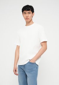 Νεαρός άνδρας που φοράει άσπρο, κοντομάνικο, υφασμάτινο πουκάμισο και ανοιχτόχρωμο μπλε τζιν, στέκεται με το ένα χέρι στην τσέπη του μπροστά σε έναν απλό φόντο.