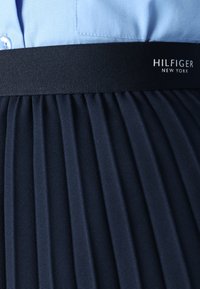 Námořnická plisovaná sukně s širokým černým pasem. Obsahuje štítek s logem "HILFIGER NEW YORK." Hladká, strukturovaná textilie vypadá pevně.