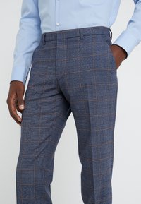 Man med blå rutiga byxor med bruna linjer och en ljusblå knapp-up skjorta, händerna i fickorna, mot vit bakgrund.