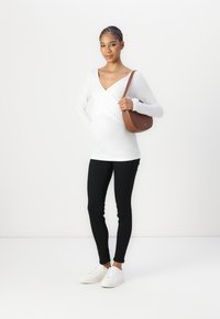 Witte lange mouwen top met een V-vormige halslijn, zwarte slim-fit broek en witte sneakers. Bruine schoudertas met een gebogen vorm.
