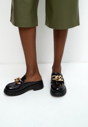 Jambes portant un pantalon court vert olive et des mocassins noirs en cuir verni avec un grand détail de chaîne dorée sur le dessus.
