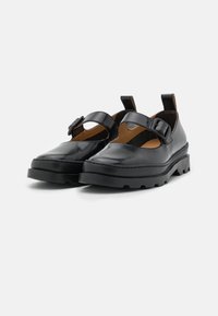 Chaussures en cuir noir avec un bout arrondi, une large bride avec boucle, une semelle épaisse et un design ajouré près de la cheville. Finition lisse et brillante.