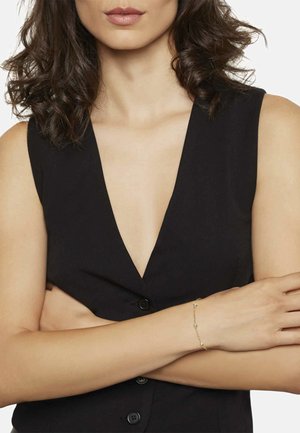 Mujer con cabello rizado oscuro, vestida con una blusa negra sin mangas y una delicada pulsera de oro con pequeños colgantes redondos en la muñeca, con los brazos cruzados.