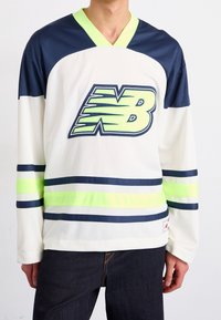 Pitkähihaisen jääkiekkopaidan väri on valkoinen, jossa on tumman sinisiä ja neonvihreitä raitoja. Paidassa on V-pääntie ja suuri vihreä "NB" logo edessä.
