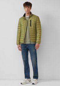 Oliwkowa puchowa kurtka ze stójką, z przodem na zamek i kieszenią na wysokości klatki piersiowej, w połączeniu z jasnym swetrem, niebieskimi dżinsami i szarymi sneakersami.