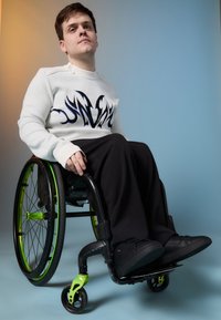White sweater with blue flame design, black pants, sitting in a black wheelchair with green accents, black shoes on footrests.