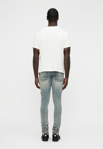 Λευκό κοντομάνικο μπλουζάκι, ανοιχτό μπλε τζιν με φθαρμένες περιοχές και πιτσιλιές μπογιάς, συνδυασμένο με μαύρα αθλητικά παπούτσια με λευκές λεπτομέρειες.