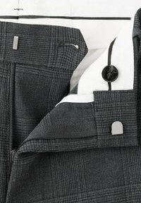 Gros plan de la ceinture d'un pantalon à carreaux gris foncé montrant une fermeture éclair, un bouton, un crochet et une barrette de fermeture, ainsi qu'une doublure intérieure blanche avec détail rayé noir.