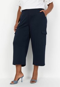Mörkblå cropped-byxor i mjukt material, med sidofickor och en avslappnad passform. Matchade med grå högklackade skor med öppen tå.