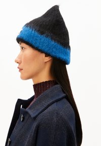 Zwarte en blauwe gebreide beanie met een puntige top. Gemaakt van zacht, textuur materiaal. Draagt bij een donkere jas en een geribde kastanjebruine trui.