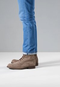 Braune Wildleder-Stiefeletten mit abgerundeten Zehen, Schnürsenkeln und einem niedrigen, geschichteten Absatz, kombiniert mit hellblauen Denim-Jeans, die am Knöchel umgeschlagen sind.