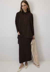 Camicia oversize marrone scuro con bottoni, abbinata a una gonna lunga coordinata. La modella tiene un tessuto color crema. Scarpe con tacco alto beige.